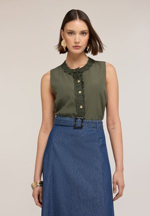 Frau trägt ärmellose olivgrüne Bluse mit gerafften Knöpfen und hoch tailliertem blauen Rock mit geflochtenem Gürtel, steht vor einfarbigem Hintergrund.