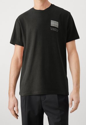 Mies pukeutunut mustaan lyhythihaiseen t-paitaan, jossa on pieni Tommy Jeans -logo ja teksti vasemmassa rinnassa, yhdistetty mustiin housuihin.
