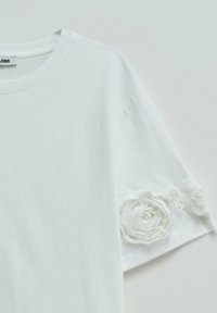 White short sleeve shirt with round neckline and two white fabric flower appliques near the hem of the right sleeve.