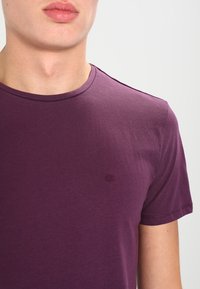 Camiseta de algodón morada con cuello redondo, mangas cortas y un sutil logo de la marca bordado en el pecho. Textura suave.