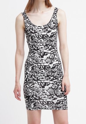 Femme portant une robe moulante sans manches à motif abstrait noir et blanc, debout devant un fond blanc uni.