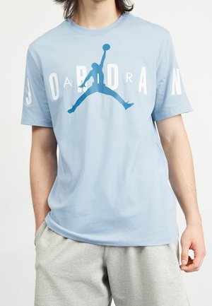 Personne portant un t-shirt bleu clair avec une silhouette de pull bleu et un texte blanc "JORDAN", associé à un short gris clair.