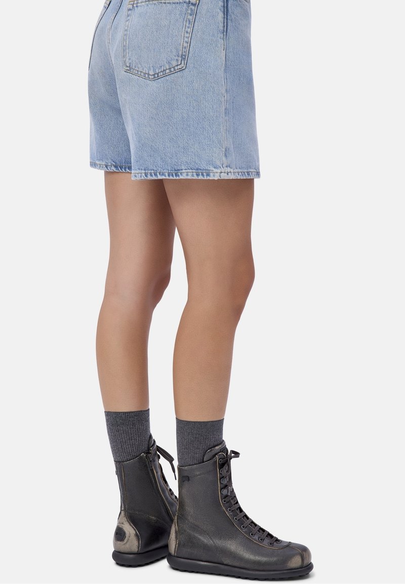 Jambes portant un short en denim bleu clair, des chaussettes gris foncé et des bottes en cuir noires montantes à lacets, debout sur un fond uni.