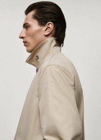 Beige trenchcoat med hög krage, med knappstängning och slät tygyta, visas från sidan mot en ljus bakgrund.