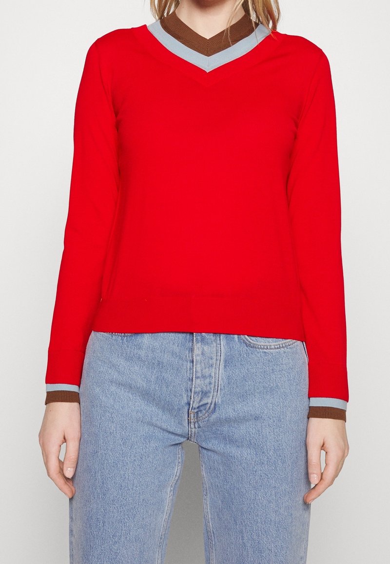 Femme portant un pull rouge à manches longues avec un col en V orné de bordures bleu clair et marron au niveau du col et des poignets, associé à un jean bleu clair.