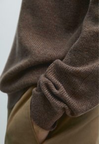 Brązowy prążkowany sweter dziany z detalem podwiniętych mankietów, wykonany z teksturowanej tkaniny, noszony na jasnym ubraniu.