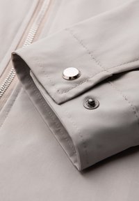 Gros plan d'un poignet de veste beige avec un bouton-pression argenté et une fermeture éclair visible sur le côté.