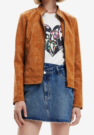 Personne portant une veste en daim beige sur un haut blanc à motif floral et une mini-jupe en jean bleu avec une fermeture éclair à l'avant.