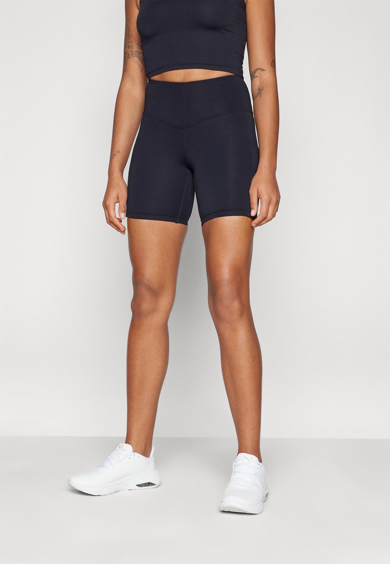 Svart atletisk crop top og matchende høyt på livet shorts laget av elastisk materiale. Hvite joggesko fullfører antrekket.