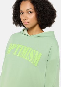 Zielona bluza z kapturem wykonana z miękkiego materiału, z napisem "OPTYMIZM" w jasnoszarych cekinowych literach. Posiada regulowany kaptur i luźny krój.