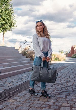 Gray duffel bag with pink polka dots and straps. Model wears a gray sweater and blue jeans, standing on cobblestone steps.