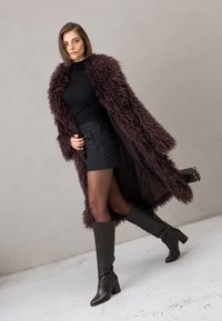 Długi, kudłaty, ciemnofioletowy płaszcz na czarnej bluzce i krótkiej spódnicy. Modelka ma na nogach brązowe, sięgające kolan skórzane kozaki i trzyma małą czarną torebkę.