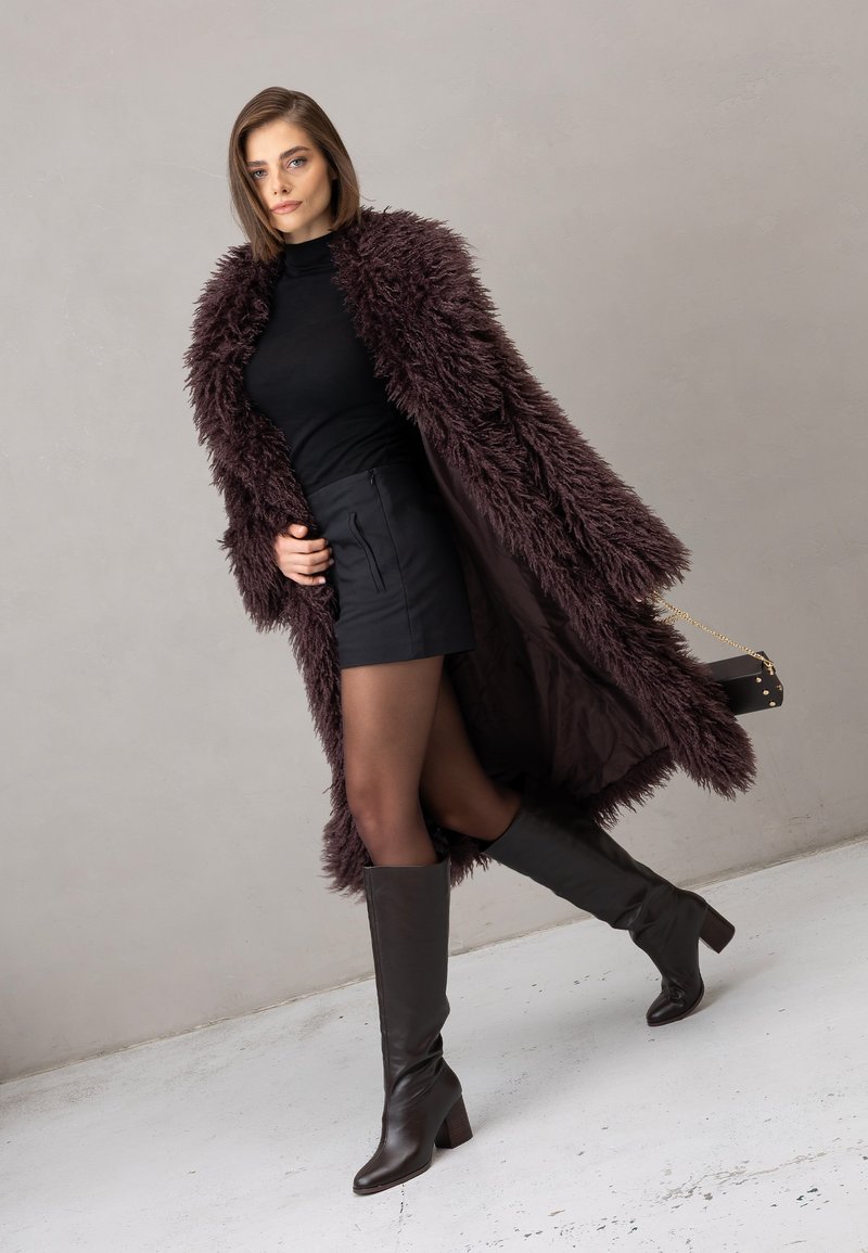 Długi, kudłaty, ciemnofioletowy płaszcz na czarnej bluzce i krótkiej spódnicy. Modelka ma na nogach brązowe, sięgające kolan skórzane kozaki i trzyma małą czarną torebkę.