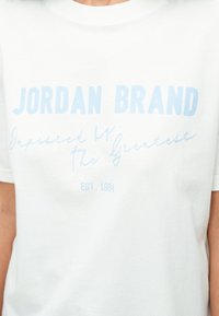 Fehér póló világoskék felirattal, amely a következőt olvassa: "JORDAN BRAND Inspired by The Greatest EST. 1984" a mellkason.
