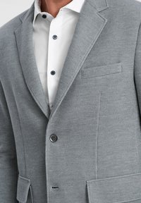 Grau strukturierter Blazer mit Reverskragen, einer Knopfverschluss und einer Brusttasche, getragen über einem weißen Hemd mit dunklen Knöpfen.