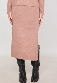 Jupe crayon en tricot de couleur rose pâle avec une texture côtelée. Longueur mi-mollet, avec un ourlet uni. Associée à des bottes noires en cuir ajustées.