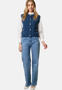 Blau gestrickte Weste mit Rippstruktur, Knopfleiste und aufgenähten Taschen über einem weißen Hemd mit Kragen, kombiniert mit hellblauen Jeans und beigen Schuhen.