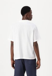 White short-sleeve linen shirt with a relaxed fit, featuring a standard collar and folded cuffs, set against a neutral background.