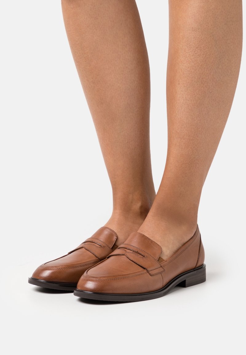 Mocassins en cuir marron avec un petit talon noir portés pieds nus sur un fond clair uni.