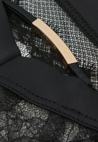Tissu en dentelle noire avec un motif texturé, comprenant une barre d'accent en métal doré et des bretelles noires lisses.
