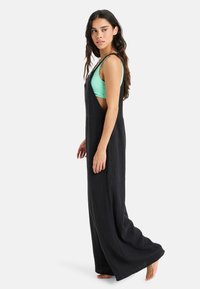 Vestido largo negro, texturizado, con tirantes anchos y cortes laterales, superpuesto sobre una blusa de color verde claro. El ajuste es relajado, llegando hasta los tobillos.