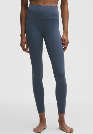 Person iført højtaljede, mørkeblå leggings stående barfodet med arme afslappet mod en ensfarvet lys baggrund.