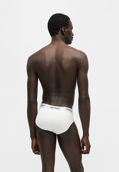 Homme debout, de dos, portant un slip blanc de la marque Off-White sur un fond clair uni.