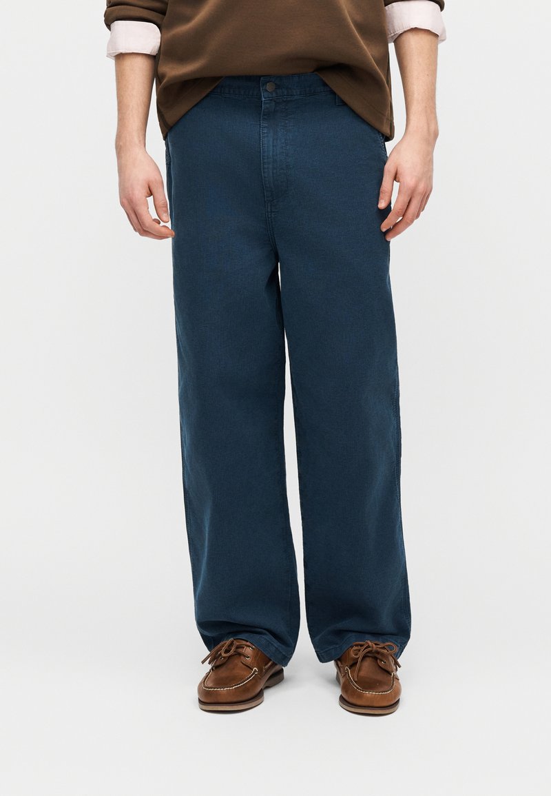Homme portant un pantalon bleu à jambes larges, des chaussures mocassins en cuir marron, et un pull marron avec les poignets de chemise blanche visibles.