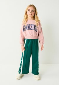 Sweatshirt rose court avec "AMAZING" en marine. Pantalons larges verts avec boutons sur le côté et rayures blanches. Baskets blanches.