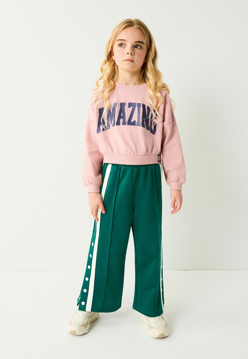 Sweatshirt rose court avec "AMAZING" en marine. Pantalons larges verts avec boutons sur le côté et rayures blanches. Baskets blanches.