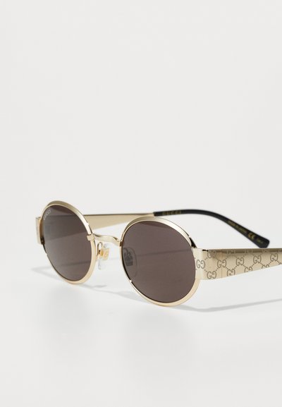 Gafas de sol en tono dorado con lentes redondos. Presentan unas patas de color beige con un diseño estampado y detalle del logo, además de lentes oscurecidos. Acentos en el puente transparente.