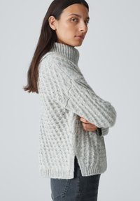 Hellgrauer gestrickter Pullover mit einem gerippten hohen Kragen, strukturiertem Muster, seitlichen Schlitzen und langen Ärmeln. Hergestellt aus weichem, bequemem Material.