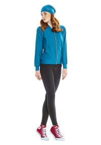 Tyrkis zip-up jacka med långa ärmar, i kombination med svarta leggings och röda höga sneakers. Modellen bär en matchande tyrkis beanie.