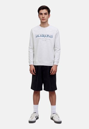 Light grey sweatshirt with blue lettering, paired with black shorts. Model stands in sneakers and white socks against a neutral background.