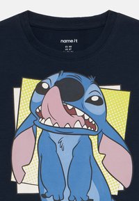 Μπλε ναυτικού μπλουζάκι με γραφικό ενός καρτούν χαρακτήρα με μεγάλα αυτιά, χαμογελαστή έκφραση και έντονα χρώματα σε φόντο με πουά.