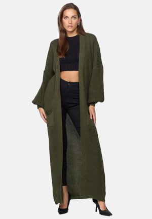 Cardigan lungo verde oliva lavorato a maglia con spalle scese, tasche frontali e design aperto, indossato sopra un crop top nero e pantaloni.