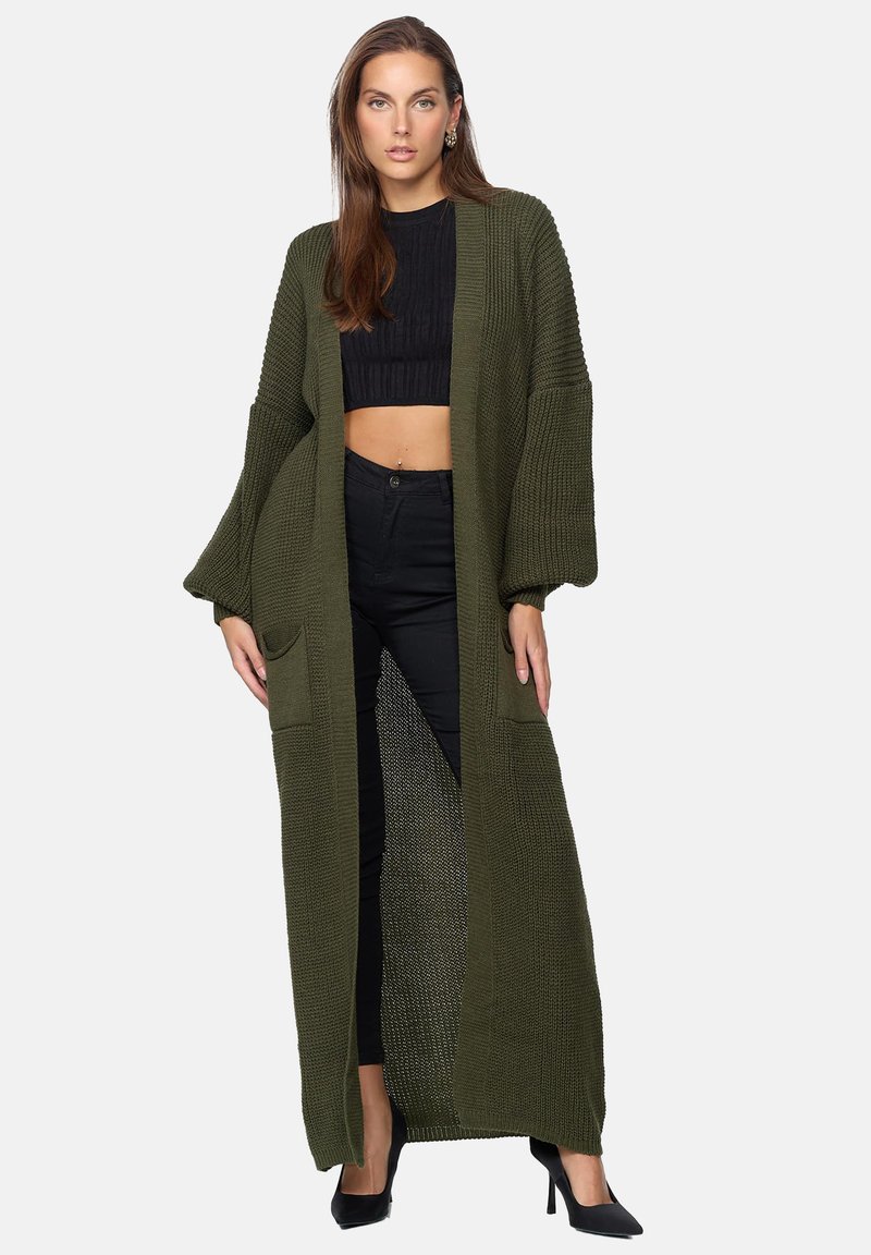 Lång olivgrön stickad cardigan med sänkta axelsömmar, framfickor och öppen frontdesign, bärs över en svart crop top och byxor.