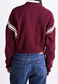 Sudadera corta color burdeos con acentos de rayas blancas en las mangas, puños y dobladillo de canalé, con un detalle de cuello en forma de V y un corte holgado.
