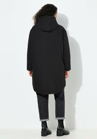 Sort hættejakke med lang kant, der har en glat tekstur og et enkelt design. Båret over mørke mønstrede bukser og chunky sorte støvler.