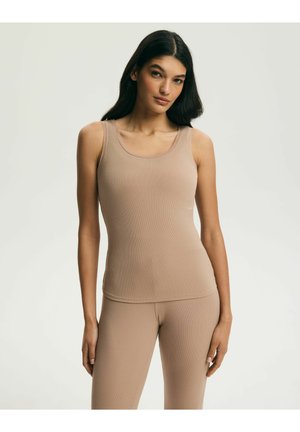 Kobieta z długimi ciemnymi włosami, ubrana w dopasowany beżowy top na ramiączkach i pasujące legginsy, stojąca na tle jednolitego jasnego tła.