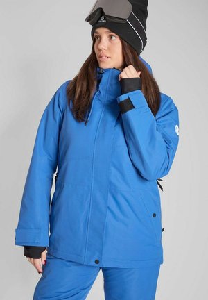 Kvinde iført en knaldblå vinterjakke og bukser med sort strikhue og skibriller løftet op på hovedet mod en grå baggrund.