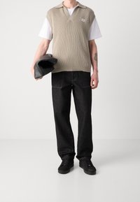 Beige gerippter ärmelloser Pullover mit V-Ausschnitt, getragen über einem weißen T-Shirt; kombiniert mit dunklen Jeans und schwarzen Sneakers; hält eine schwarze Tasche.