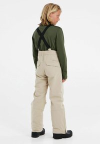 Beige Schneehosen mit verstellbaren schwarzen Trägern, geradem Schnitt und Reißverschlussöffnungen an den Knöcheln. Tragen über einem grünen Langarmoberteil.