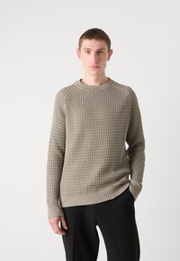 Beige gestrickter Pullover mit gerippter Textur, Rundhalsausschnitt und langen Ärmeln. Kombiniert mit schwarzen Hosen, die einen lockeren Schnitt und dezente Details aufweisen.