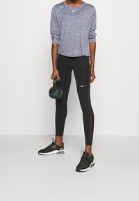 Dlouhý rukáv Nike, strukturovaný tmavě modrý top; černé elastické legíny s síťovanými detaily; drží černou kettlebell; černé sportovní boty.