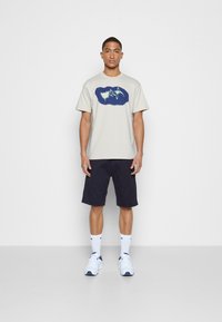 T-shirt cinza claro de manga curta com um design gráfico azul, combinado com calções azul-marinho e ténis brancos, de pé contra um fundo simples.