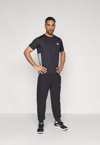 Sort atletiktrøje med hvid Adidas-logo på brystet, kombineret med sorte joggingbukser og sorte sneakers med hvide sidestriber.