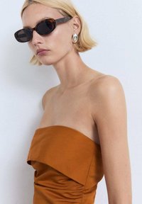 Bruine strapless jurk met gerimpelde details. Schildpadbril en grote metalen oorbellen complementeren de look. Over het algemeen een soepele textuur.