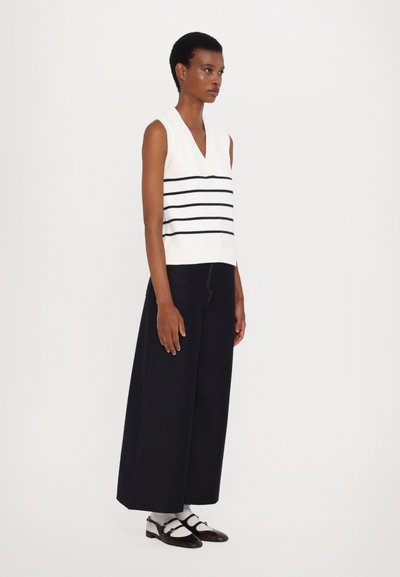 Weißer ärmelloser Pullover mit marineblauen horizontalen Streifen und V-Ausschnitt. Kombiniert mit weit geschnittenen dunklen Hosen und dunkelbraunen Lederschuhen.