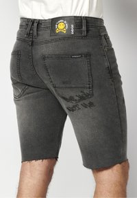 Shorts en denim noir avec ourlets effilochés, poches arrière, patch jaune avec smiley et texte "Don't Worry Be Happy" sur la ceinture.